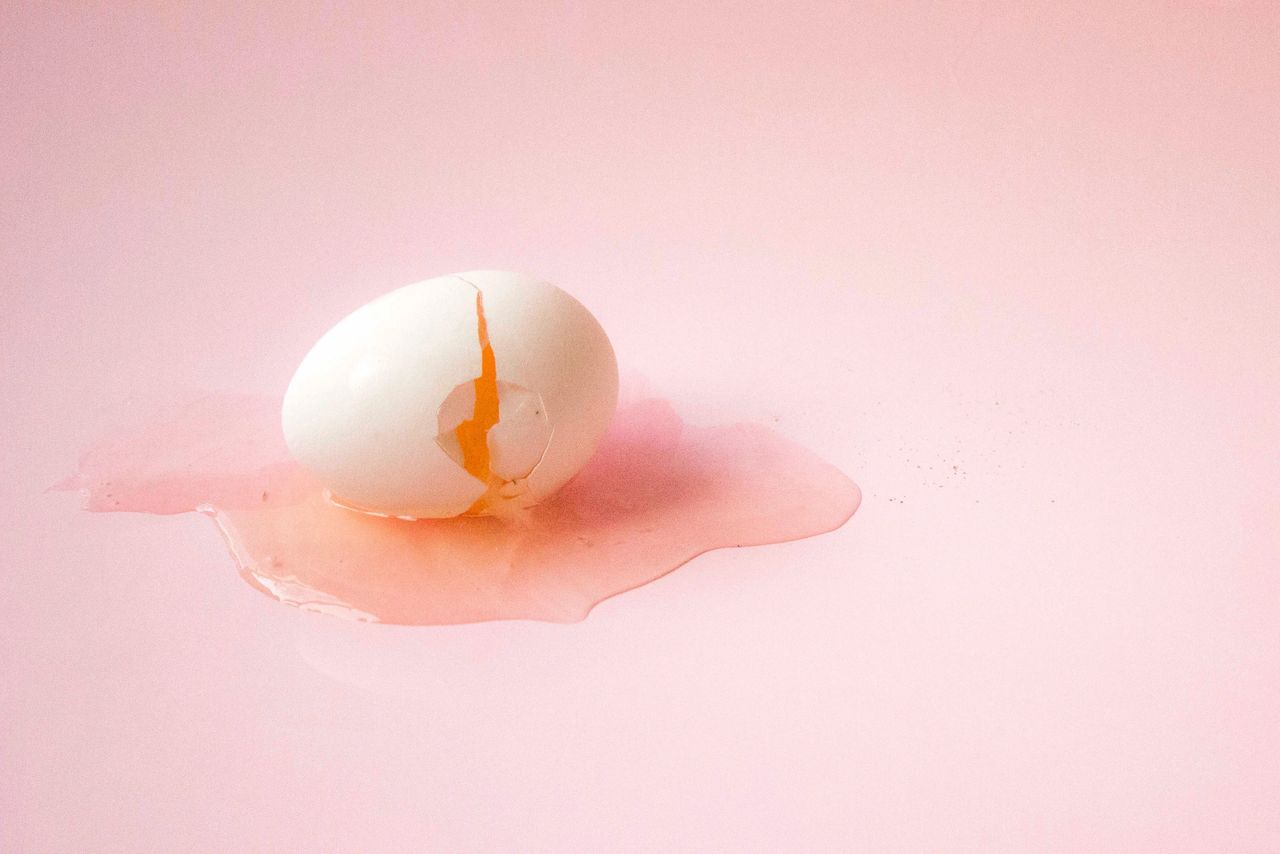 A cracked egg leaking yolk on a pink background. Photo by Melani Sosa on Unsplash.